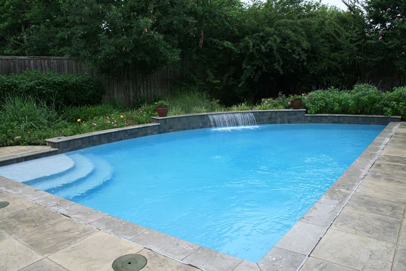 This Denton, Texas swimming pool design features a tile veneered multi-level raised beam with a large sheer descent waterfall, a honed travertine coping, stamped concrete decking and superblue aggregate interior finish.