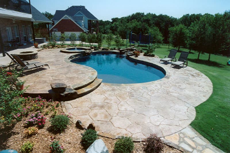 This Argyle, Texas swimming pool features stamped concrete decking, stone steps, Oklahoma flagstone coping, a boulder veneered raised beam, an elevated spa, a boulder waterfall and Tahoe blue aggregate interior finish