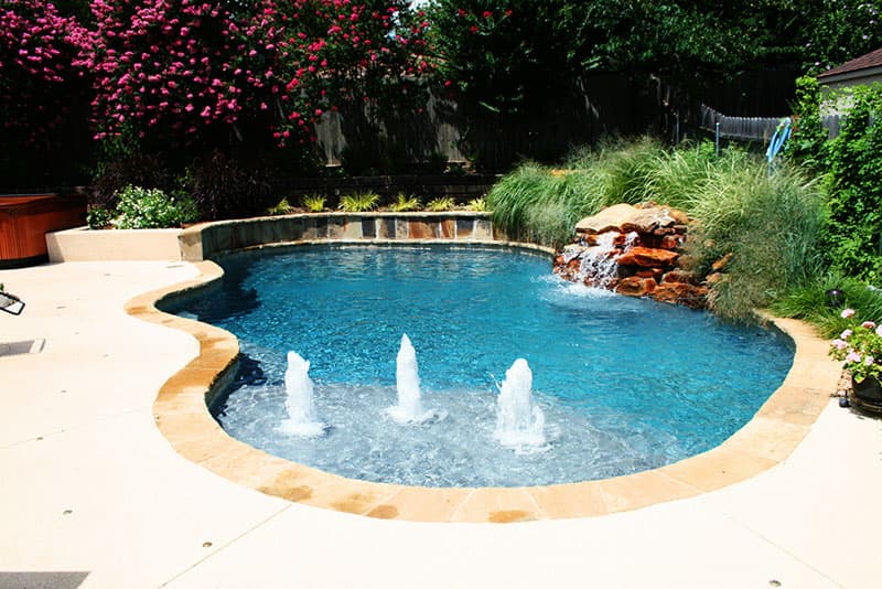 This Denton, Texas swimming pool features a boulders waterfall tucked into the lush landscaping, Oklahoma flagstone coping and veneered raised beam, decorative concrete spray- decking, a large tanning ledge with fountains and pebble finish interior.