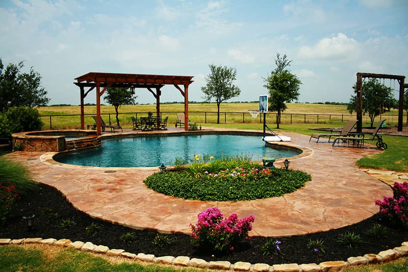 This Sanger, Texas swimming pool exudes relaxation from the moment you hear the faint whisper of the tiered spillway on the old hickory stone veneered spa to the sight of the handcrafted shade structure. This families backyard dreamscape also features stamped concrete decking, Oklahoma flagstone coping, Midnight blue aggregate interior finish, a water-basketball goal and large landscaping planters.