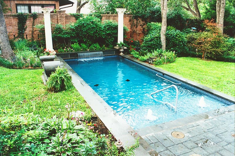 This Denton, Texas award-winning swimming pool design features two large Grecian style columns atop a Pennsylvania veneered raised beam that contains a 36 inch stainless steel sheer descent waterfall, Pennsylvania blue-stone coping and matching stamped concrete decking, a large tanning ledge with fountains, powder coated water exercise bar and handrail, lush landscaping and a midnight blue aggregate interior finish.