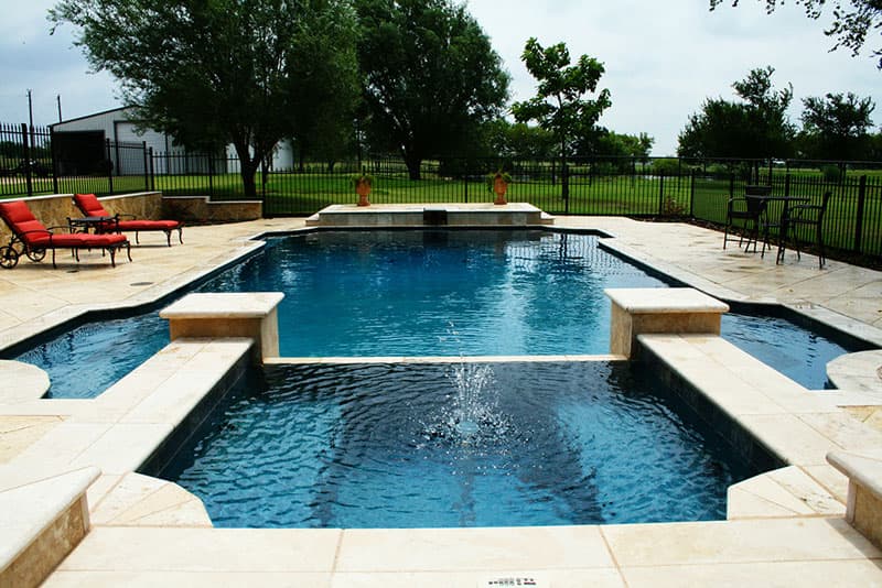 This Sanger, Texas swimming pool not only compliments to the beauty of the wide open property but addresses the elevation changes with an Oklahoma flagstone veneered retaining wall capped with travertine. The elevated spa features matching honed travertine columns, a tiered fountain and spillway. The travertine coping outlines the main pool’s competing entry steps and benches to the recessed diving board of the travertine tile veneer of the raised beam where one may prepare to plunge into the deep aquatic color of the Mediterranean blue aggregate interior finish as others relax on the sun bleached stamped concrete decking