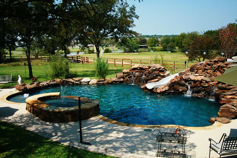 This Denton, Texas swimming pool features a mid-sized boulder waterfall and a massive boulder waterfall which supports a custom water-slide and multiple splash points, a stone staircase that leads to the slide entry, accent boulders, Oklahoma flagstone coping, concrete spray-decking, an elevated spa with flat stone spillway and Mediterranean blue aggregate finish interior.