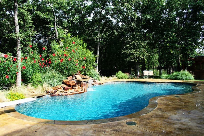 This Flower Mound, Texas diving pool features a large grotto waterfall, Oklahoma flagstone coping, stain concrete decking, diving board, a large tanning ledge and Tahoe blue aggregate interior finish.