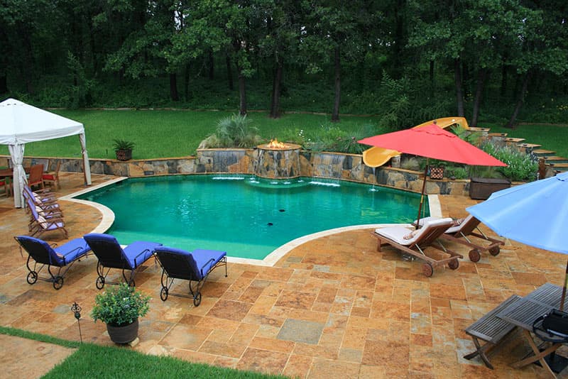 This Argyle, Texas swimming pool features travertine decking with contrasting travertine coping, a large irregular flagstone raised beam that is centered by a firepit with scupper water features, to one side it connects to a matching retaining wall and on the other stone steps lead to a custom water-slide.