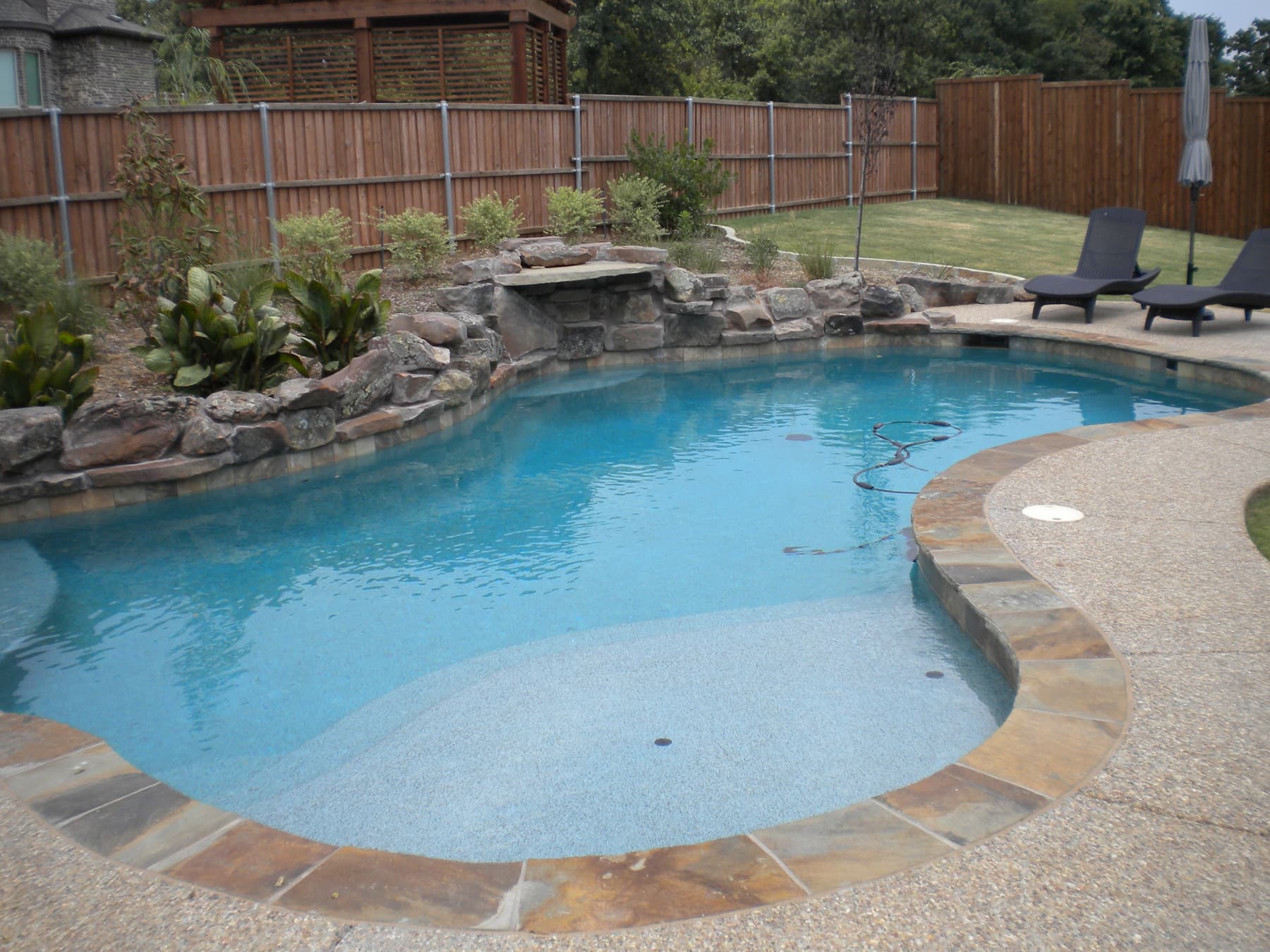 The natural boulder waterfall serves as a backdrop to this Denton County area pool. Exposed aggregate decking and a random colored Oklahoma flagstone envelop the Tahoe blue waters created by the interior finish.