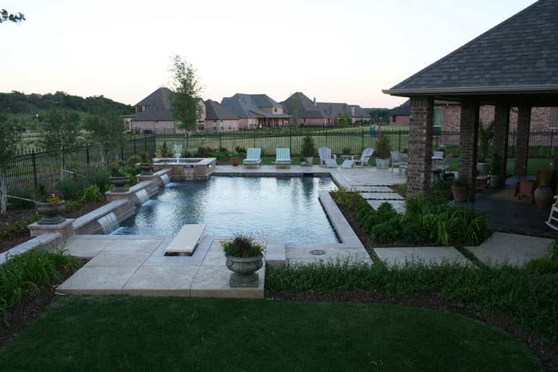 This Country Club Village project incorporates an elevated spa with stainless steel spillways and a 3-tiered fountain which blends into a varying height raised wall with 3 sheer descent waterfalls trimmed in a tumbled travertine. A 6′ recessed diving board serves as the launching point into the blue granite pebblesheen finish.