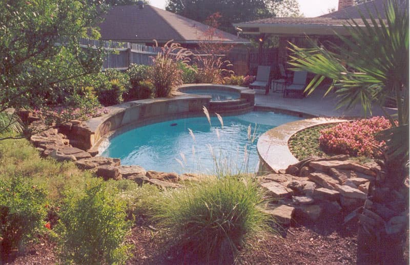 A moss boulder waterfall serves as the backdrop for this South Denton, Texas swimming pool. A Tahoe blue aggregate pool surface accentuates the Oklahoma flagstone used as coping and veneer for the raised beam and the elevated spa as it protrudes from the hillside into the washed aggregate decking.