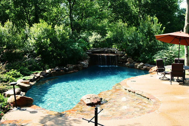The acid stained decking of this Denton, Texas swimming pool lends way to the all stone tanning ledge where you can sit in awe of the massive grotto and boulders snuggled into the green foliage of the hillside.