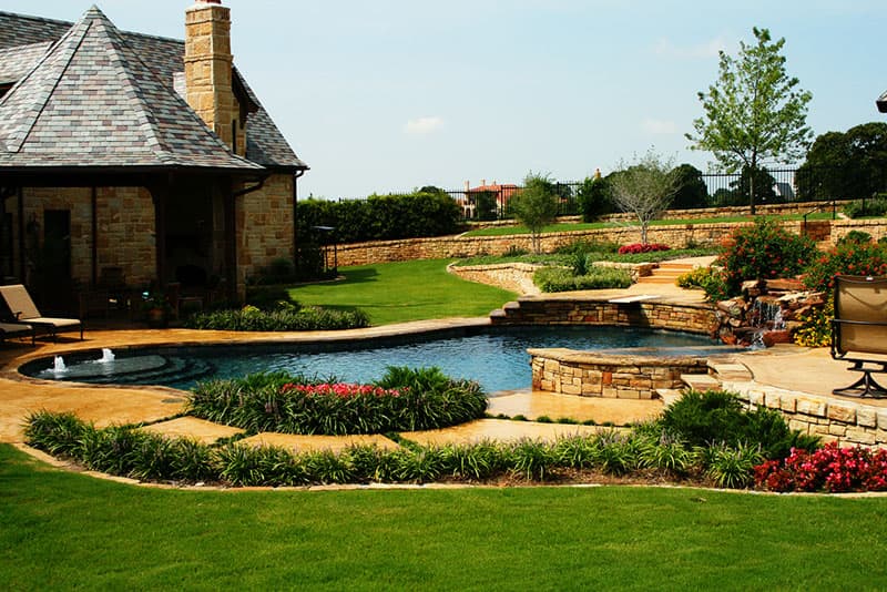 This manicured environment is located in the Vaquero community of Westlake, Texas. The raised beams Oklahoma stone match the existing walls as the large boulder waterfalls splash into the midnight blue aggregate finish. The colorful landscaping lends to the stained concrete decking as it leads you to the elevated diving board.