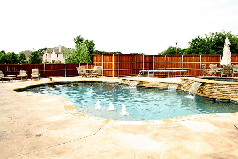 Three sheer descent waterfalls spill from the Oklahoma stone veneer of the raised wall and spa into the deep blue water accented by tanning fountains and stamped concrete decking of this Denton swimming pool.