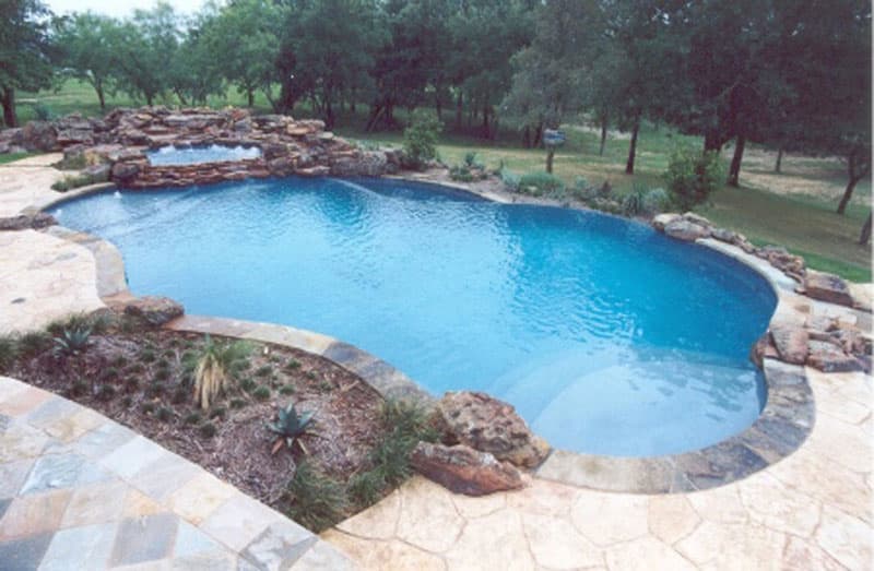 Located in Little Elm, Texas this negative edge swimming pool was crafted using a mixture of Oklahoma flagstone and moss rock boulders. The dual tanning ledges oppose each other. The boulder spa spills into the main pool as a water slide hidden by boulders exits into the lower basin, Planters and multi-level stamped decking engulf this waterscape.