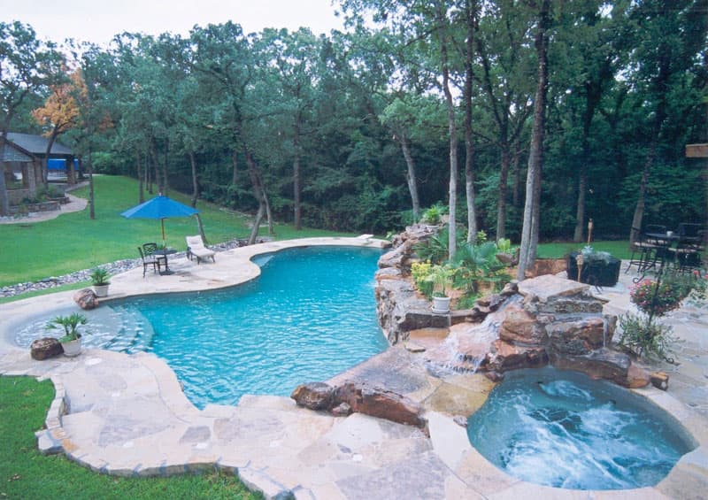 The large boulder waterfall serves as the backdrop of this Denton, Texas pool as it emerges seamlessly from the hillside and begins at the grotto spillway of the elevated spa and ends in the lower level diving area. Stone decking and Oklahoma flagstone lead you to the entrance of the Tahoe blue pebble finish of the tanning ledge.