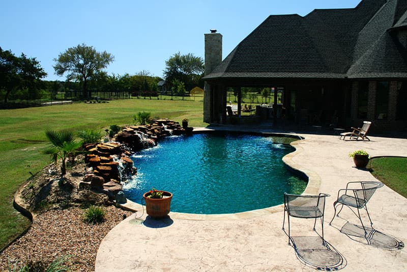 This Argyle, Texas swimming pool was designed with a rustic style in mind; moss rock boulders waterfalls, Tumbled travertine coping, stamped concrete decking, midnight blue pebble finish interior, large tanning ledge and elevated spa are used to achieve this look.