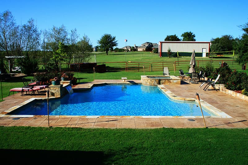 This Aubrey, Texas diving pool design features stamped concrete decking, stone retaining walls, a raised beam with a sheer descent waterfall, Oklahoma coping, elevated spa, diving board and Tahoe blue aggregate finish.