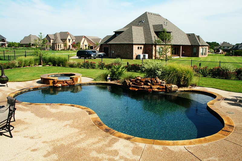 Washed aggregate decking leads you to this Argyle, Texas residential swimming pool, from the Oklahoma veneer of the elevated spa to the natural stone waterfall as it spills onto the pebble finish of the tanning ledge it is truly an environment built for relaxation.