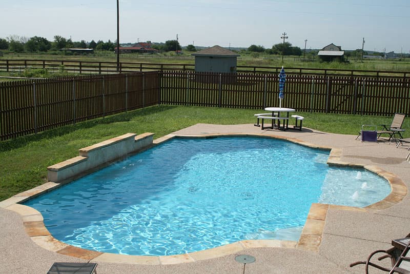 This Argyle, Texas swimming pool features a 3 tiered raised wall with a sheer descent waterfall, tanning ledge with water features, buff colored spray-decking and superblue aggregate interior finish.