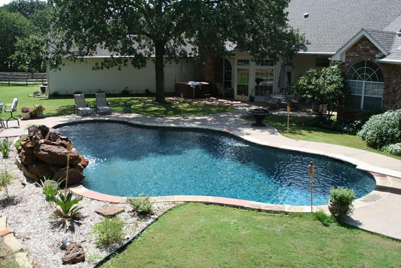 This Argyle, Texas swimming pool features a diving board, a boulders waterfall, concrete spray-decking, a large tanning ledge with water features and a Tahoe blue aggregate finish interior.